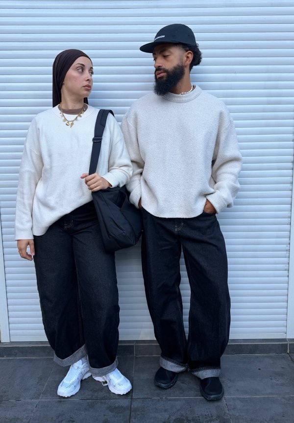 Dve osebi v belih puloverjih in črnih hlačah stojita drug ob drugem ob belem kovinskem roloju in se gledata.