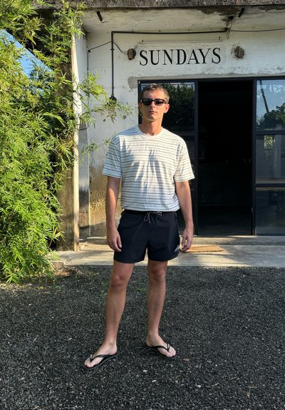 Camiseta de manga corta a rayas en beige claro y gris oscuro, combinada con pantalones cortos de baño negros y chanclas negras, de pie al aire libre.