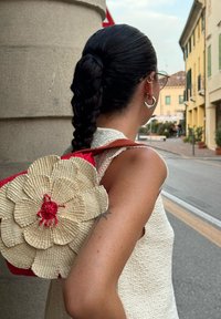 Woven straw σακίδιο με μεγάλο μπεζ σχέδιο λουλουδιού και κόκκινες λεπτομέρειες. Η τσάντα διαθέτει υφασμάτινη επιφάνεια και ρυθμιζόμενους ιμάντες ώμου.