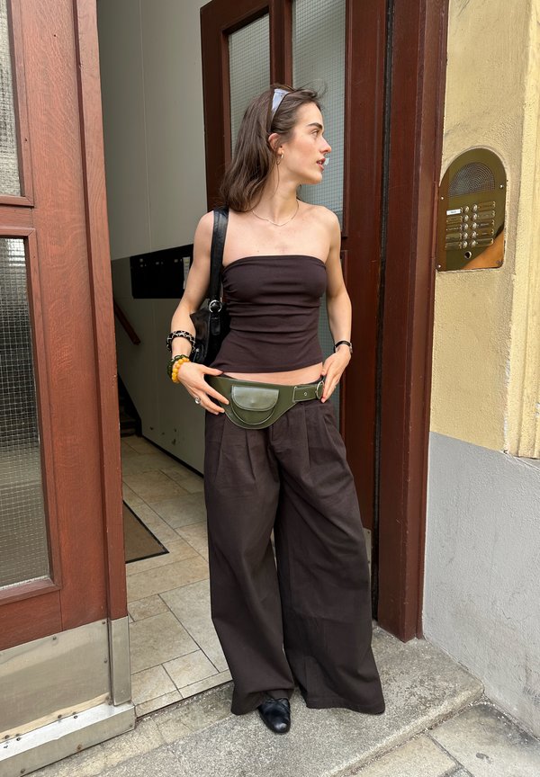 Jeune femme en haut noir sans bretelles et pantalon large ajustant un sac ceinture vert, debout dans un cadre de porte près d’un panneau d’interphone.