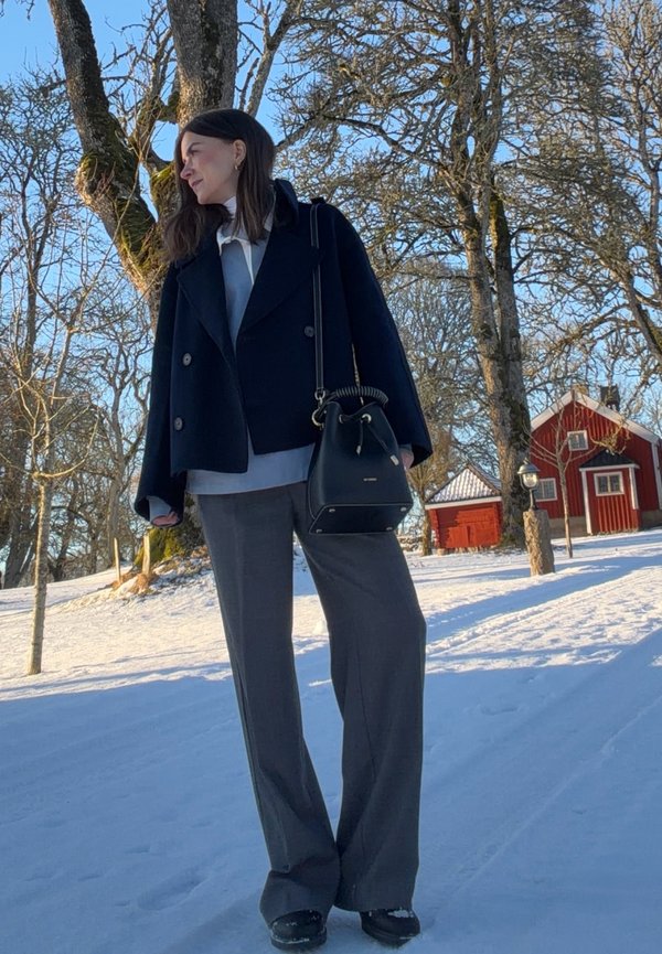 Donna con cappotto nero e pantaloni grigi in piedi in un cortile innevato con alberi spogli e case di legno rosse sotto un cielo azzurro limpido.