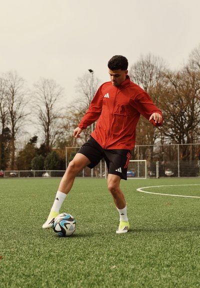 Joven con chaqueta roja y pantalones cortos negros controlando un balón de fútbol en un campo de césped con postes de portería y árboles al fondo.