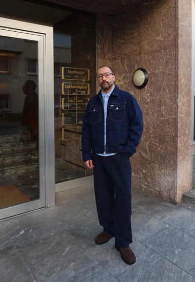 Hombre con gafas, chaqueta azul marino y pantalones, de pie frente a un edificio con puertas de cristal y carteles comerciales en una pared de piedra marrón.