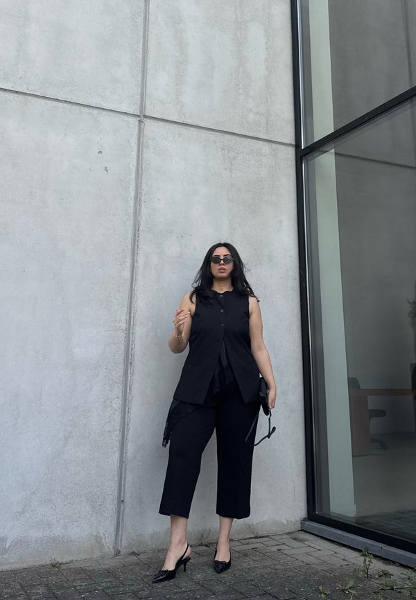 Frau trägt schwarze ärmellose Weste, schwarze verkürzte Hose und Sonnenbrille, steht auf dem Gehsteig neben einer Betonmauer und einem Glasfenster.