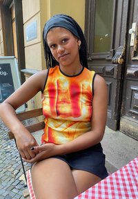 Top sem mangas com um gráfico de mãos em laranja brilhante, amarelo e vermelho, combinado com uma saia preta. Cabelo trançado e uma faixa escura na cabeça.