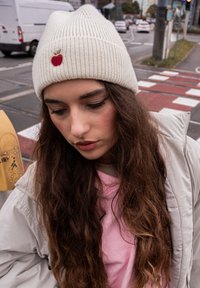 Crème ribbelgebreide beanie met een kleine rode appelbrodering; gedragen met een lichtgrijze jas over een roze top. Lange, golvende bruine haren zichtbaar.