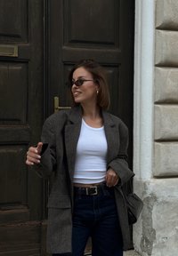 Grå ullblandad oversized blazer över en vit kortärmad linne, i kombination med hög midja mörkblå jeans och svarta accessoarer. Strukturerad bakgrund.