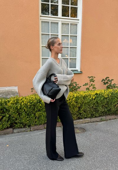 Suéter gris de gran tamaño con ribetes, bolso estructurado negro y pantalones anchos negros. Texturas suaves y colores neutros. Entorno al aire libre.