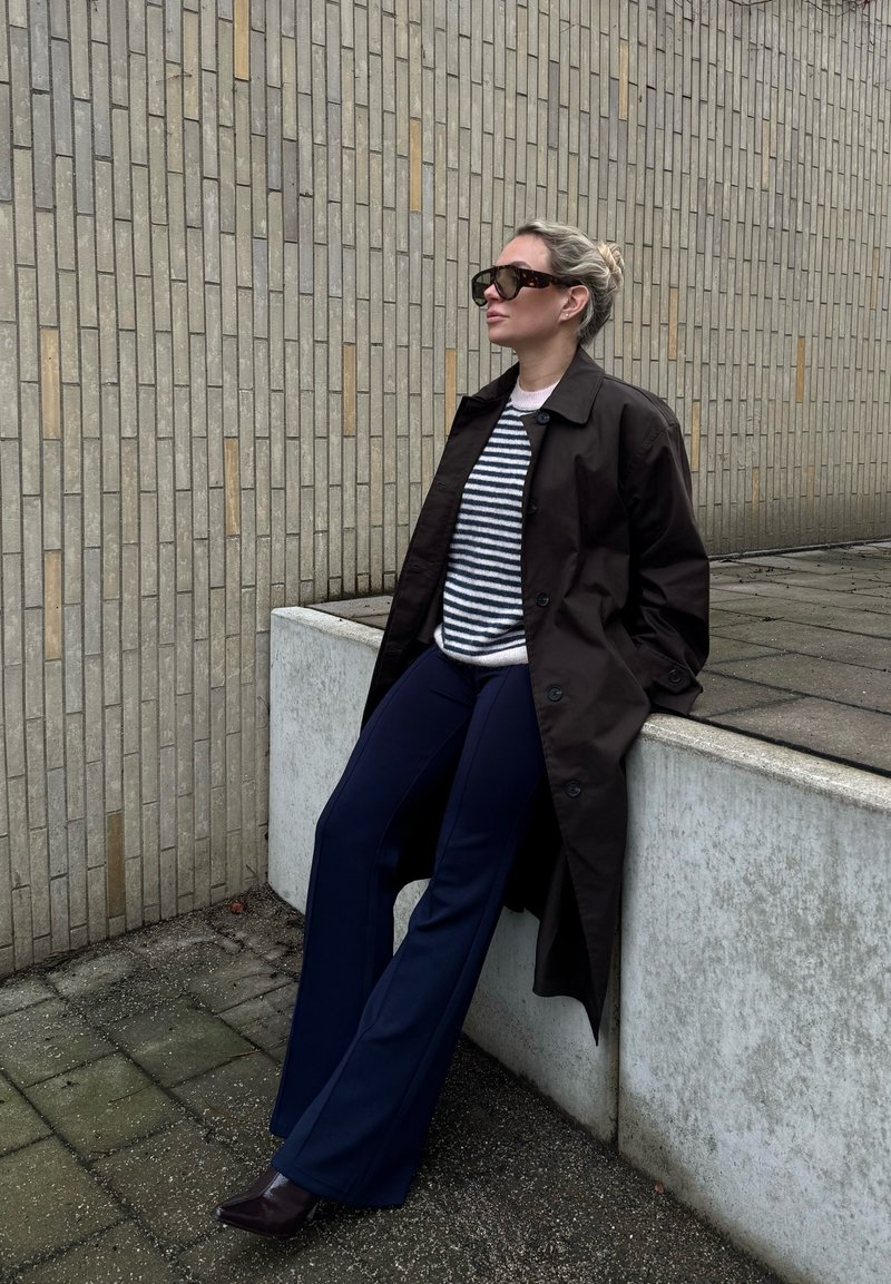 Femme aux cheveux blonds attachés en chignon, portant des lunettes de soleil, un pull rayé, un manteau foncé et un pantalon bleu marine, s'appuie sur un rebord en béton près d'un mur en briques à l'extérieur.