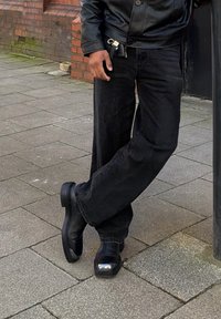 Personne portant une veste en cuir noire, un pantalon noir et des bottes noires à bout métallique, debout sur un trottoir pavé près d'un mur en briques.