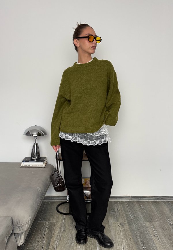 Pull en maille vert olive, bord en dentelle blanche en dessous, pantalon noir, chaussures noires et lunettes de soleil teintées jaunes. Style moderne avec des textures superposées.