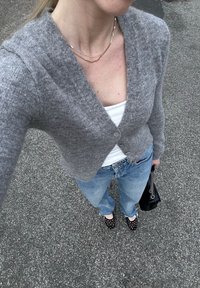 Cardigan grigio in maglia con scollatura a V, chiusura con bottoni, abbinato a una canottiera bianca e jeans azzurri chiari. Scarpe nere con pois e una borsa nera.