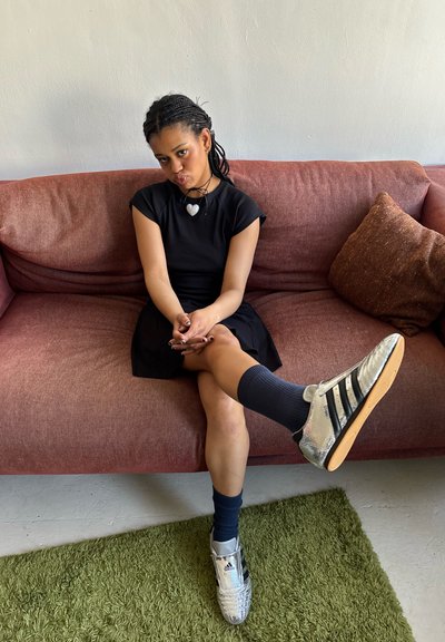 Vestido negro, zapatillas plateadas brillantes con tres rayas negras, calcetines azul oscuro y un collar con un colgante de corazón. Sentada en un sofá rojizo.