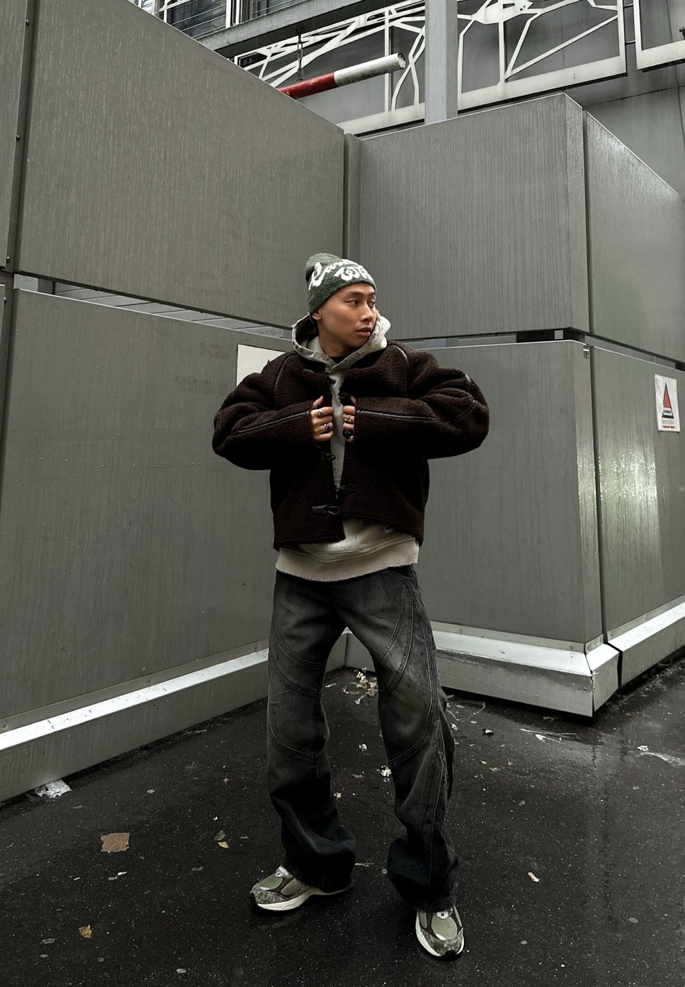 Ruda, tekstūruota striukė dėvima virš pilko džemperio su gobtuvu, kartu su laisvais pilkais džinsais ir sidabriniais sportiniais bateliais. Aksesuarai apima žalią megztą kepurę.