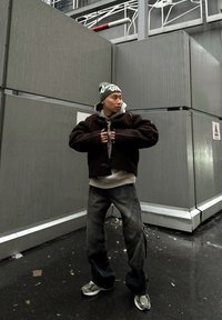 Ruda, tekstūruota striukė dėvima virš pilko džemperio su gobtuvu, kartu su laisvais pilkais džinsais ir sidabriniais sportiniais bateliais. Aksesuarai apima žalią megztą kepurę.