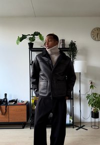 Veste en cuir noire avec deux poches avant, fermeture à boutons et un col doux, superposée à un col roulé texturé clair. Pantalon noir.