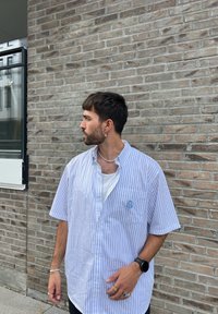 Košile s krátkým rukávem, zapínaná na knoflíky, s modrobílými vertikálními pruhy, s kapsou na levé straně hrudi a vyšitým logem, přetažená přes bílé tílko.