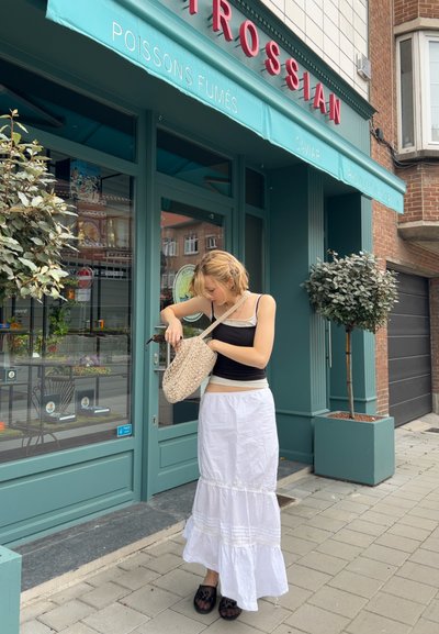 Bolso de paja tejido, camiseta negra sin mangas, falda en capas de color crema y beige con volantes. Escena de acera, edificio verde con toldo, plantas en macetas.