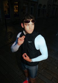 Veste noire sans manches avec logo Nike, chemise à manches longues blanche, short gris et chaussures roses. Un homme ajuste sa veste dans un environnement humide.