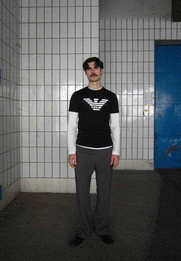 T-shirt preta com logotipo branco, sobreposta a uma camisa de manga longa branca. Calças cinzentas largas. Figura isolada contra uma parede azulejada.