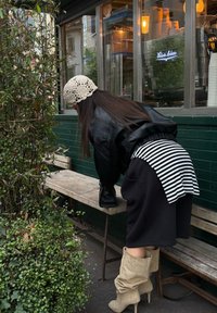 Casaco de couro preto, sobreposição de tecido às riscas, saia preta e botas bege até ao joelho. Vegetação ao redor e banco de madeira visíveis.