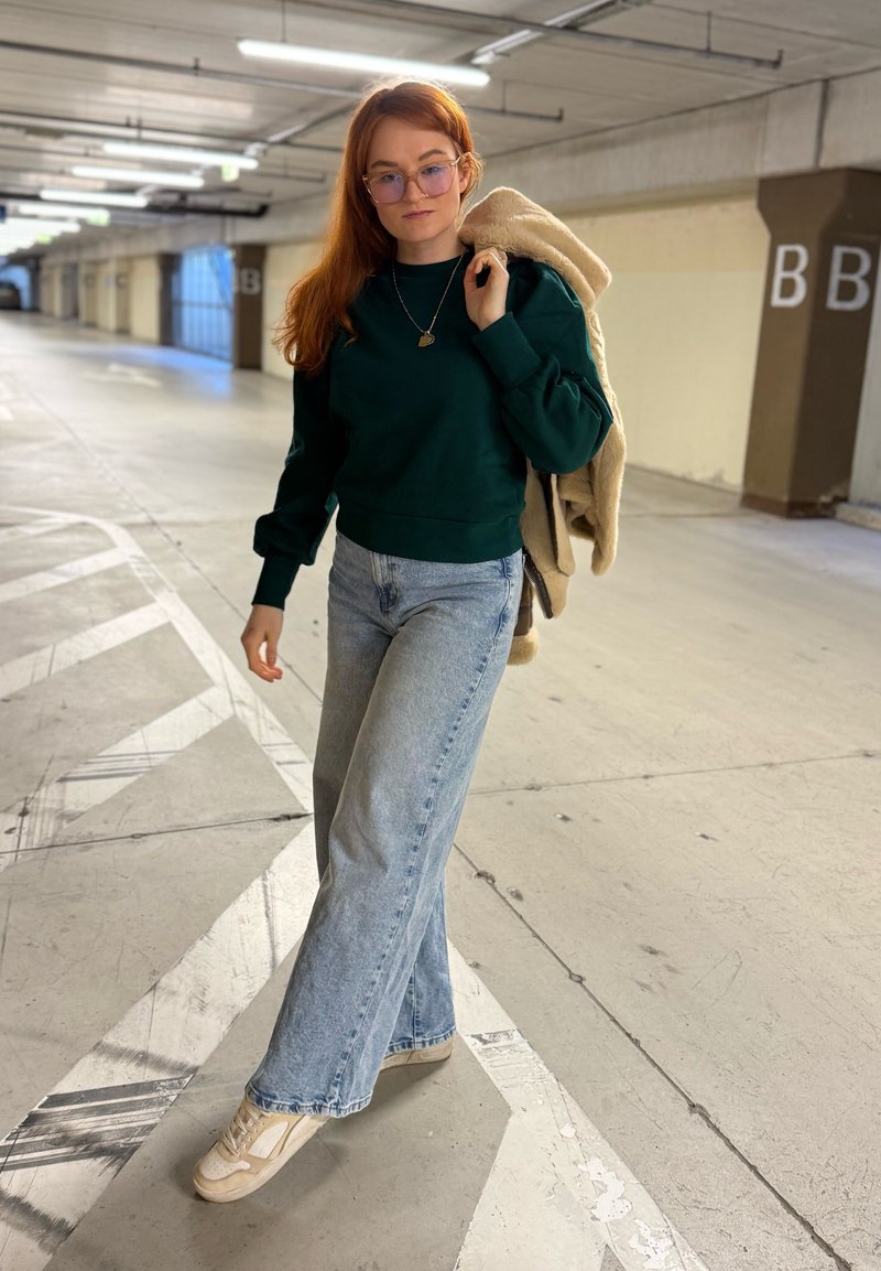 Sweat-shirt en coton vert, jean large bleu clair, baskets blanches. Porte une veste beige. En arrière-plan, un parking avec des marquages.