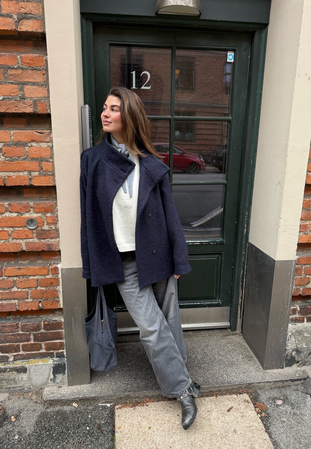 Temno modra plašč iz mešanice volne z ovratnikom typu šala, oblečen preko svetlega puloverja. Sivke široke hlače in črni gležnarji. Temno siva torba.