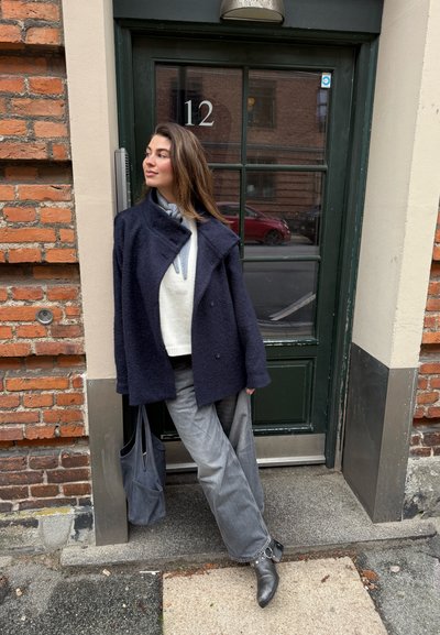 Abrigo azul marino de mezcla de lana con cuello chal, superpuesto sobre un suéter ligero. Pantalones anchos grises y botines negros. Bolso tote gris oscuro.