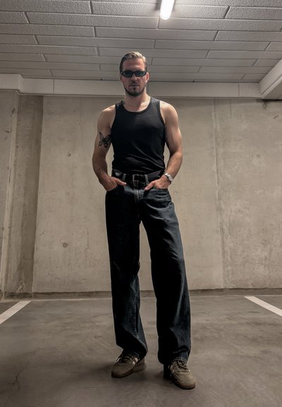 Hombre con camiseta sin mangas negra, vaqueros oscuros, gafas de sol y zapatillas de deporte, de pie con las manos en los bolsillos en un aparcamiento de hormigón.