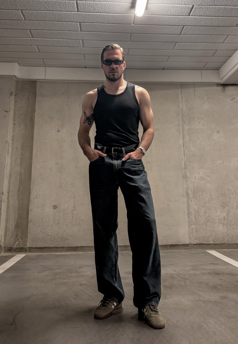 Homme portant un débardeur noir, un jean foncé, des lunettes de soleil et des baskets, debout les mains dans les poches dans un parking en béton.