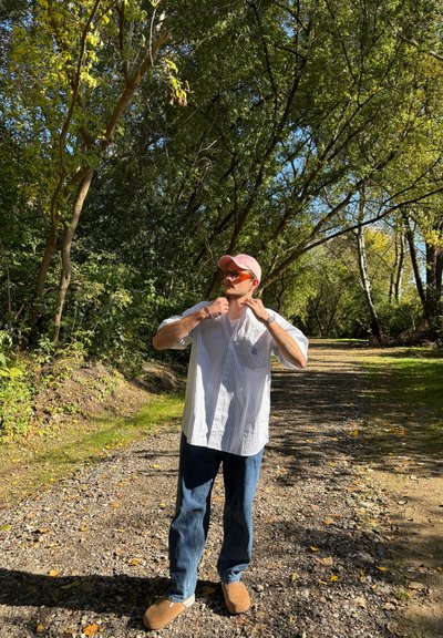 Camisa de manga corta a rayas, azul claro y blanca; jeans azules; pantuflas marrones; gorra rosa; gafas de sol redondas; de pie en un camino de grava en un área forestal.