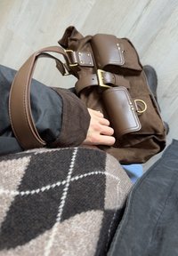 Mano che tiene in mano una borsa in suede e pelle marrone con cintura dorata, indossa una giacca nera e un maglione con motivo argyle, in piedi su un pavimento di legno chiaro.