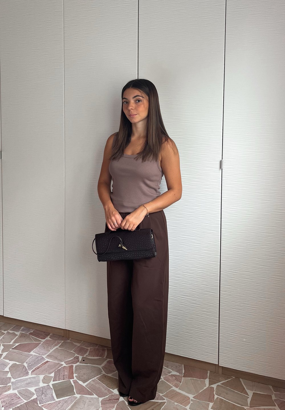 Bruine wijde broek gecombineerd met een aansluitend mauve hemdje. Een zwarte textuur clutch wordt voor zich gehouden. De achtergrond toont lichtgekleurde keukenkastjes.