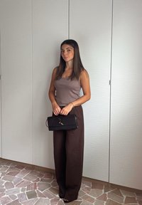 Bruine wijde broek gecombineerd met een aansluitend mauve hemdje. Een zwarte textuur clutch wordt voor zich gehouden. De achtergrond toont lichtgekleurde keukenkastjes.