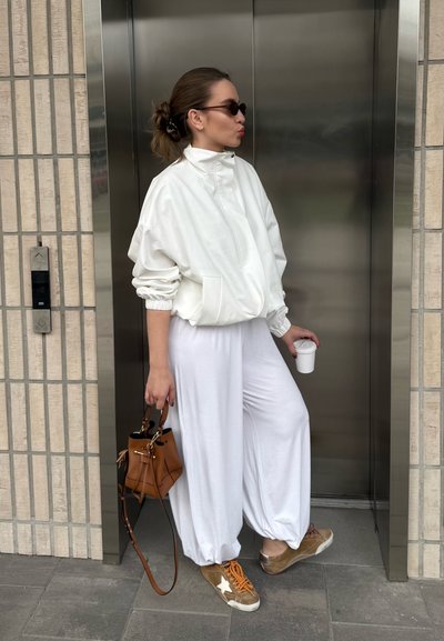 Mujer vestida con chaqueta y pantalones blancos de gran tamaño, zapatillas marrones, que sostiene un bolso marrón y una taza blanca, de pie junto a una puerta de ascensor cerrada.