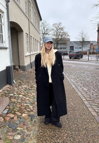 Largo abrigo negro, sudadera crema, pantalones negros y una gorra de béisbol azul clara. De pie en una calle adoquinada cerca de edificios.
