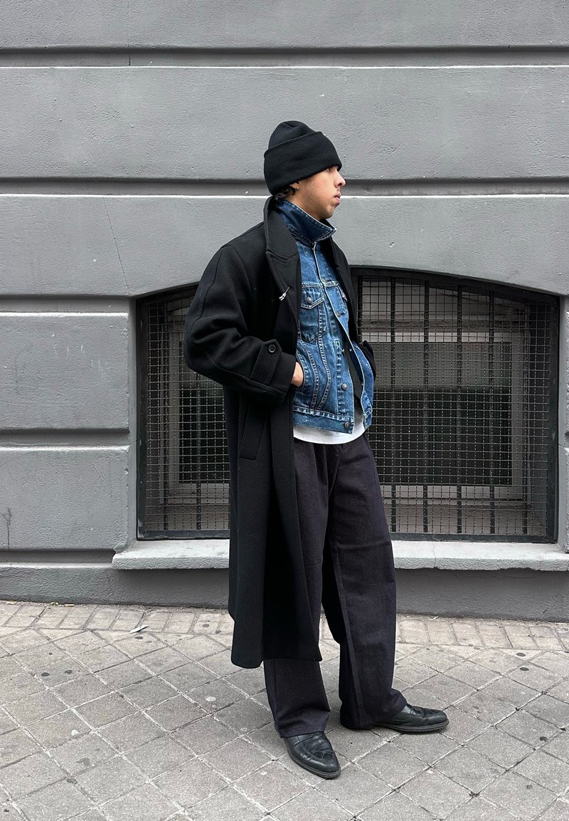 Jeune personne portant un bonnet noir, un manteau noir superposé et une veste en jean bleu, debout de profil contre un mur gris avec une fenêtre à barreaux.