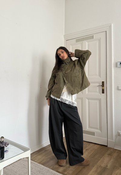 Chaqueta verde oliva con cuello alto, superpuesta sobre una camiseta blanca con bordes de encaje. Pantalones negros de pierna ancha combinados con zapatos color marrón claro. Ambiente minimalista.