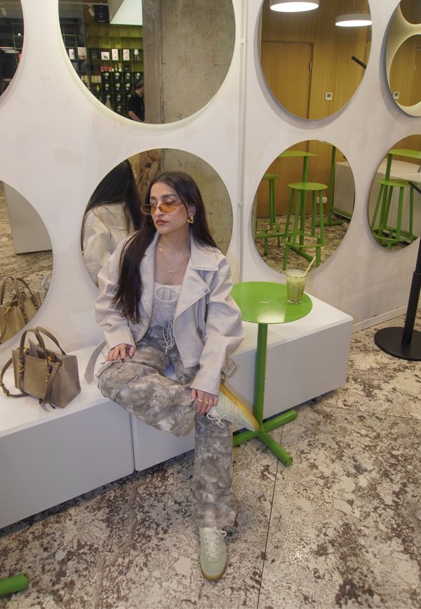 Grijze en beige oversized jas, tie-dye broek, groene sneakers. Zittend naast een groene bijzettafel met een drankje, tegen ronde spiegels.