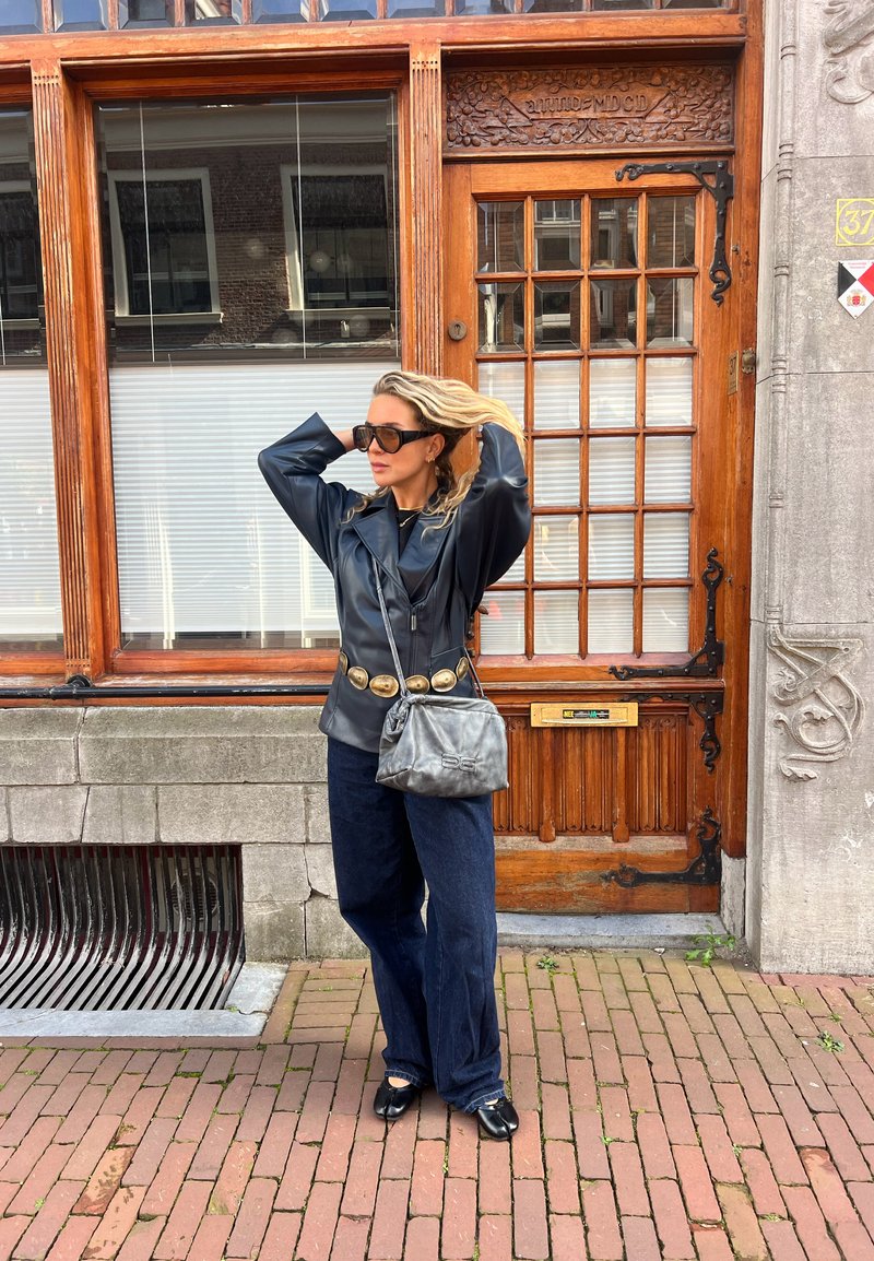 Giubbotto di pelle nera con accenti dorati, pantaloni di denim scuro larghi e una borsa grigia. Abbinato a occhiali da sole neri di grandi dimensioni, in piedi davanti a una porta di legno.
