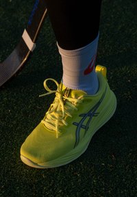 Zapatilla deportiva de color amarillo brillante con una parte superior texturizada, detalles en azul y cordones planos. Usada con un calcetín blanco que lleva un logo rojo.