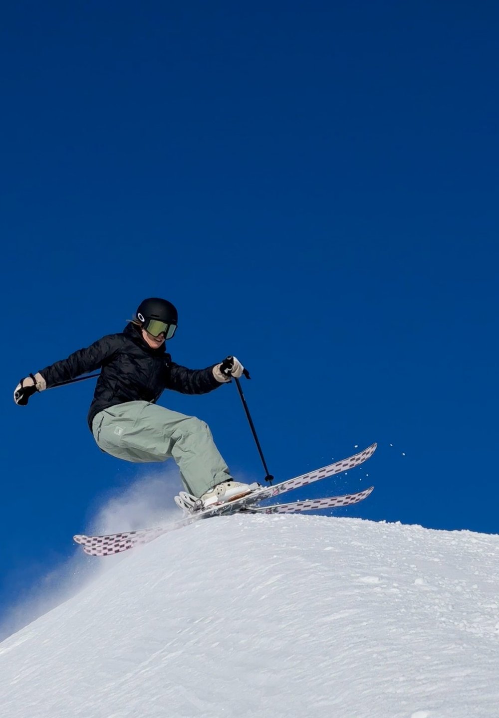 O equipamento de esqui apresenta um esquiador com uma jaqueta preta e calças verde-claro, utilizando esquis brancos com detalhes em rosa, numa encosta nevada contra um céu azul.