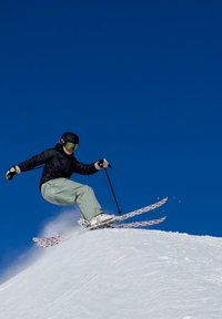 O equipamento de esqui apresenta um esquiador com uma jaqueta preta e calças verde-claro, utilizando esquis brancos com detalhes em rosa, numa encosta nevada contra um céu azul.