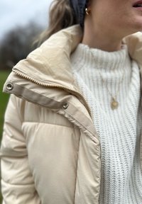 Casaco puffer bege claro com colarinho alto e botões de pressão, combinado com um suéter de malha branca grosso. Colar de ouro visível.