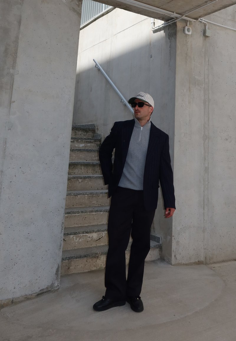 Ein Mann mit Sonnenbrille, grauem Reißverschluss-Pullover, dunklem Blazer, Hose und Kappe steht nahe Betonstufen und Wänden in einem industriellen Umfeld.
