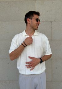 Polo shirt bianco lavorato a maglia con un motivo verticale testurizzato, maniche corte e colletto a punta, abbinato a pantaloni grigi chiari.