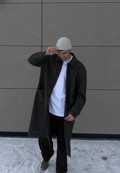 Hombre con gorro gris, abrigo largo oscuro, camisa blanca y pantalones negros de pie sobre nieve frente a una pared de azulejos grises, ajustándose el sombrero.