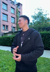 Melnā sporta jaka ar rāvējslēdzēju, priekšējā kabatā un ar logotipu uz krūšu daļas, valkāta virs tumša grafiskā T-kreklu, āra vidē.