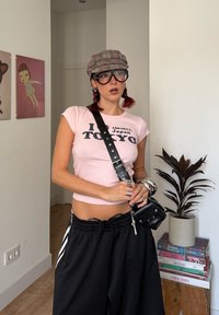 Tricou roz cu mâneci scurte cu textul „I 160-0011, Japan TOKYO”, asortat cu pantaloni largi negri. Accesorizat cu o șapcă în carouri și ochelari mari.