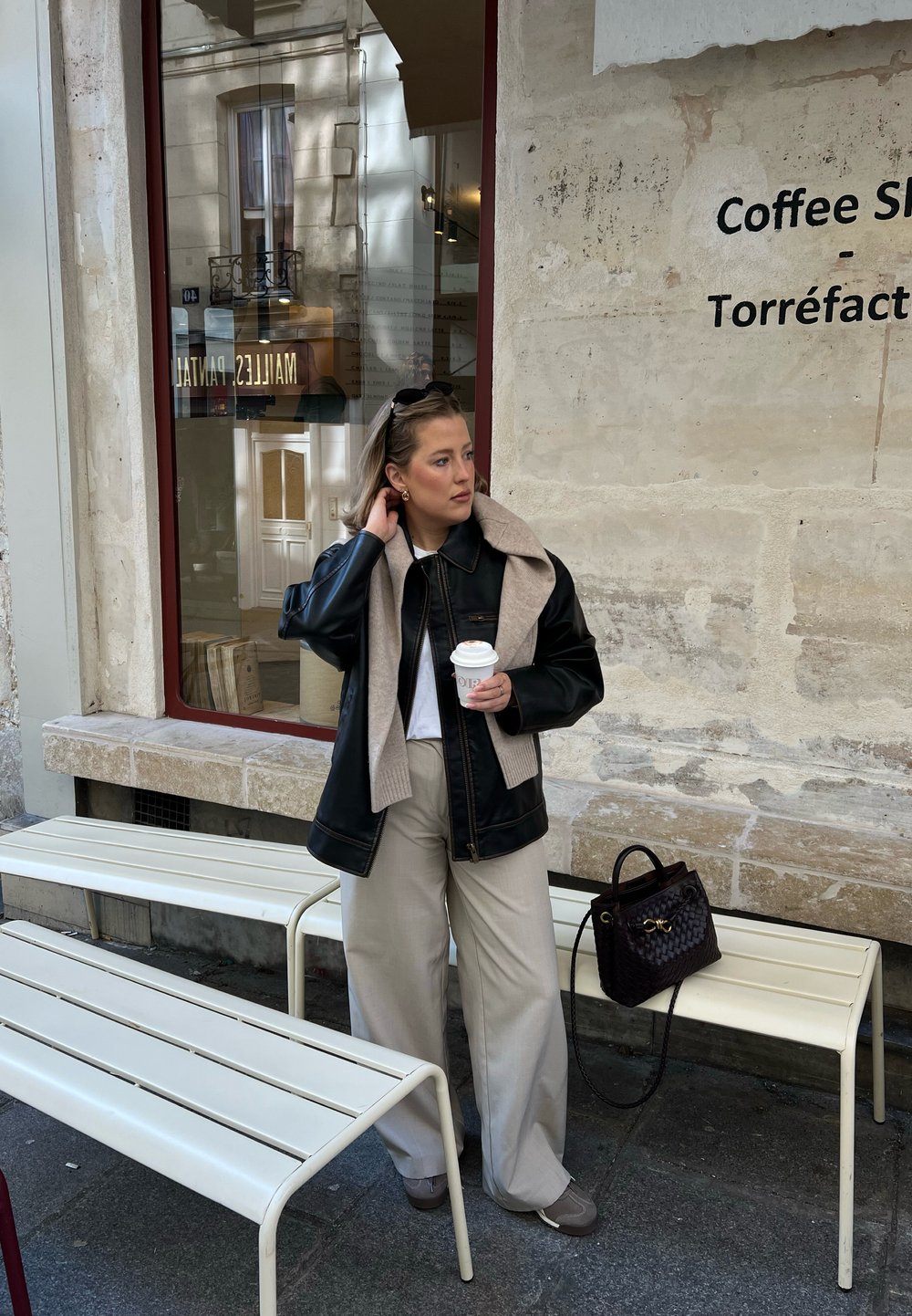 Giacca di pelle nera con sciarpa grigia, t-shirt bianca e pantaloni larghi beige chiaro. Tiene in mano una tazza di caffè, mentre sta in piedi accanto a panchine bianche.